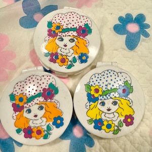 Puede incluir: Tres espejos compactos blancos con tapa abatible. Cada espejo presenta una ilustración colorida de una niña con cabello rubio que lleva un sombrero con lunares y flores. La cara de la niña está rodeada de flores.