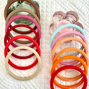 May include: A collection of colorful bangles in various shades, including red, pink, orange, and beige. The bangles are arranged near two decorative glass dishes. The bangles are made of a smooth, glossy material.