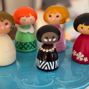 May include: A collection of six vintage doll figurines with rounded heads and painted faces. The dolls have different colored hair and dresses, including red, green, and black. The dolls are displayed on a light blue, decorative cake stand.