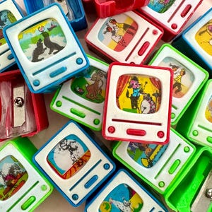 以下が含まれることがあります： 青、緑、赤、白など、さまざまな色のヴィンテージテレビ型の鉛筆削りのコレクション。各鉛筆削りは、前面に異なる漫画の画像が描かれています。