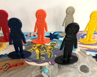 May include: A set of six astronaut figurines, each in a different color: red, blue, orange, gray, black, and yellow. The figurines are standing on oval bases and are made of plastic.