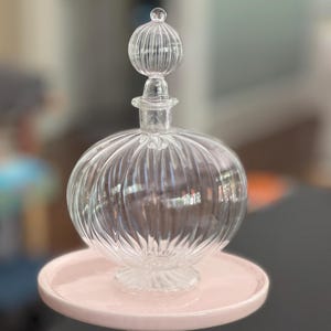May include: A clear glass decanter with a rounded, ribbed body and a matching spherical, ribbed stopper. The decanter features a short neck and a small, circular base, showcasing a transparent design with vertical ridges throughout the glass.