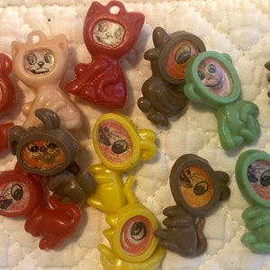 Peut inclure: Une collection de 12 breloques en plastique en forme de chat avec des corps et des visages de différentes couleurs. Les breloques ont un trou en haut pour être accrochées. Les chats sont dans différentes poses, y compris assis, debout et couchés.