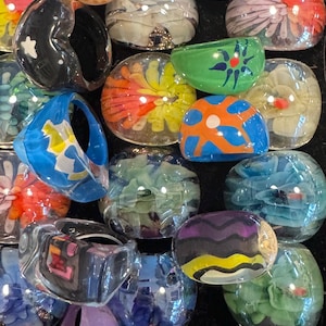 May include: A collection of colorful, handcrafted glass rings and bowls. The rings and bowls feature various designs, including floral patterns, geometric shapes, and abstract art. Colors include blue, orange, yellow, green, purple, and black.