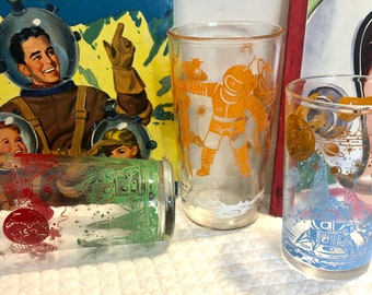 May include: Three vintage drinking glasses with space-themed designs. The glasses are clear with orange, green, and blue designs. The designs include astronauts, rockets, and planets.
