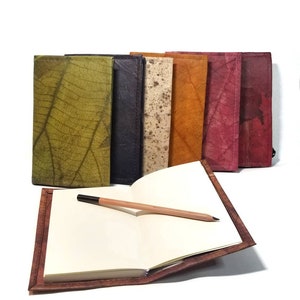 May include: A set of six colorful leather-bound journals with a blank journal open and a pencil on the page. The journals are made from recycled materials and have a unique, natural look.
