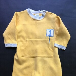 May include: A yellow baby onesie with white trim and a blue patch with the number "1" on it.