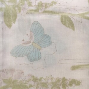 May include: A light-colored fabric with a repeating pattern of butterflies and floral elements. The butterfly is light blue with a pink body and yellow accents. The floral elements are in shades of green and pink.
