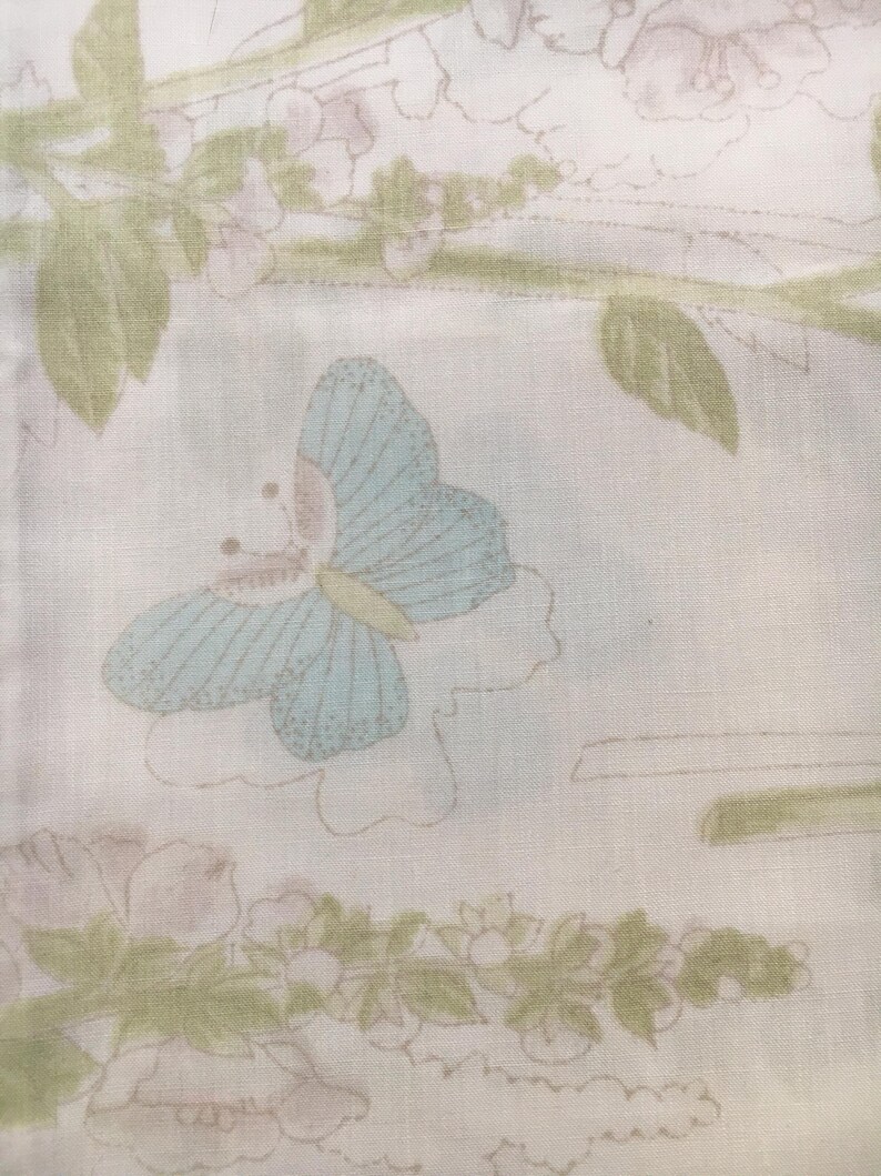 May include: A light-colored fabric with a repeating pattern of butterflies and floral elements. The butterfly is light blue with a pink body and yellow accents. The floral elements are in shades of green and pink.