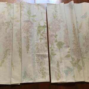 May include: Two folded white fabric panels with a floral pattern. The design features pink and green flowers with green leaves and light blue butterflies. The panels are likely curtains or decorative fabric.