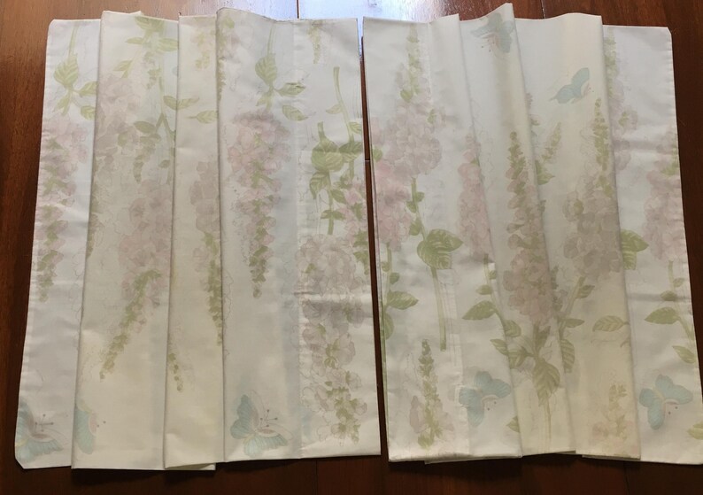 May include: Two folded white fabric panels with a floral pattern. The design features pink and green flowers with green leaves and light blue butterflies. The panels are likely curtains or decorative fabric.