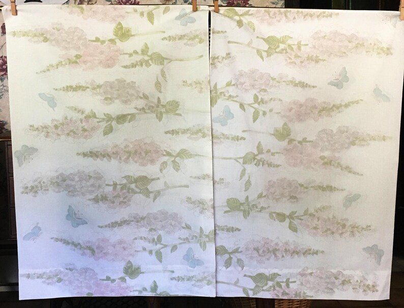 May include: Two white fabric panels with a repeating pattern of pink and lavender floral branches and light blue butterflies. The fabric is held up by wooden clothespins.