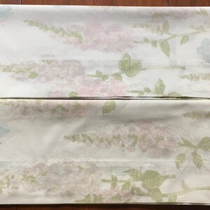 May include: Two white bed sheets with a floral pattern. The design features pink and lavender flowers, green leaves, and light blue butterflies. The sheets are folded and laid flat.