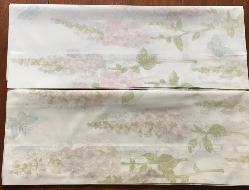May include: Two white bed sheets with a floral pattern. The design features pink and lavender flowers, green leaves, and light blue butterflies. The sheets are folded and laid flat.