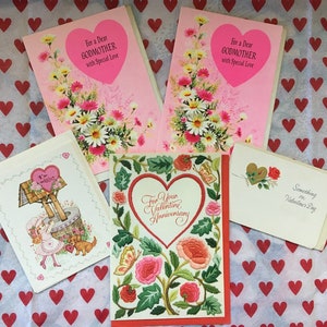 May include: A collection of vintage greeting cards with floral and heart designs. The cards include messages like "For a Dear Godmother with Special Love" and "For Your Valentine Anniversary". The cards are on a red heart-patterned background.