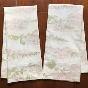 May include: Two white floral patterned towels. The towels feature a repeating design of pink flowers and green leaves. The towels are folded and displayed on a wooden surface. The floral pattern is soft and delicate.