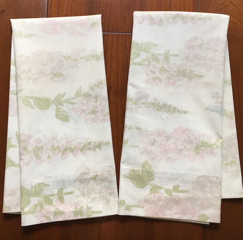May include: Two white floral patterned towels. The towels feature a repeating design of pink flowers and green leaves. The towels are folded and displayed on a wooden surface. The floral pattern is soft and delicate.