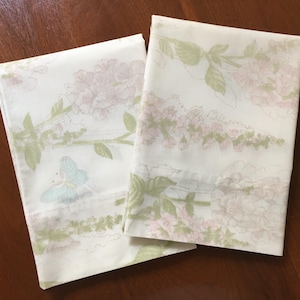 May include: Two folded white pillowcases with a floral pattern. The design features pink flowers, green leaves, and a light blue butterfly. The pillowcases are on a dark brown wooden surface.