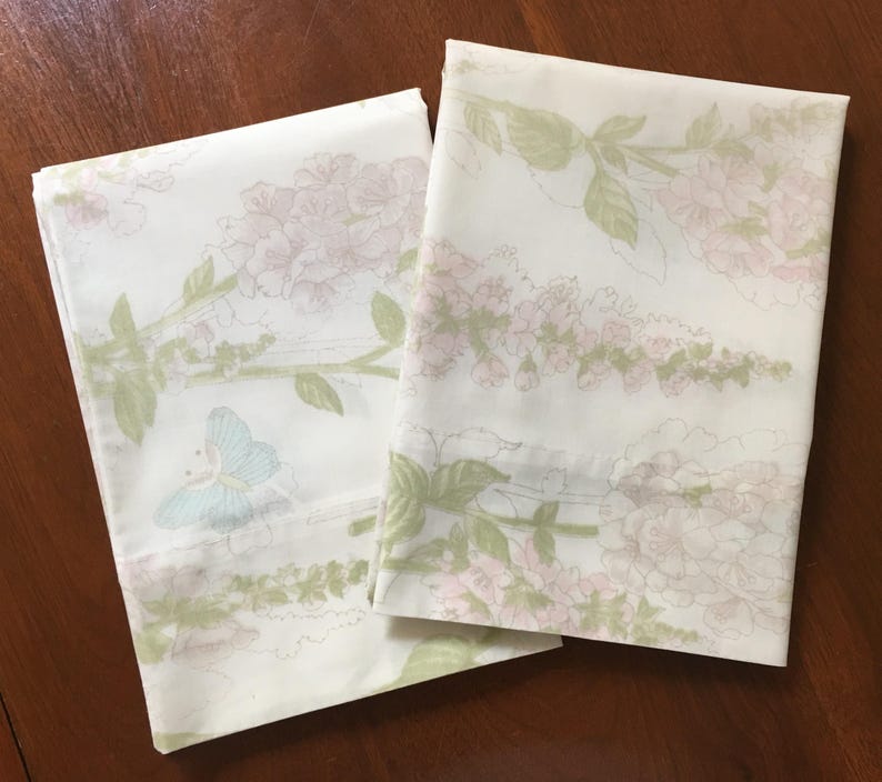 May include: Two folded white pillowcases with a floral pattern. The design features pink flowers, green leaves, and a light blue butterfly. The pillowcases are on a dark brown wooden surface.