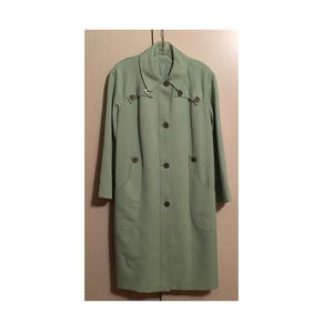 May include: A light sage green long-sleeve coat featuring a pointed collar, a central button-down closure, and two side pockets. The garment includes decorative button accents on the chest area and long sleeves, providing a structured silhouette.