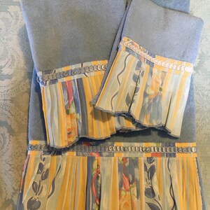 May include: Set of three blue bath towels with a yellow, blue, and white floral print trim. The towels have a ruffled edge and a white ribbon.