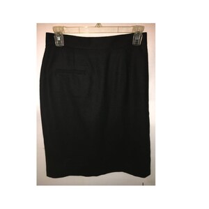 May include: A black pencil skirt hanging on a white background. The skirt is made of a solid black fabric and has a classic, tailored design. It features a back vent and a single pocket detail.