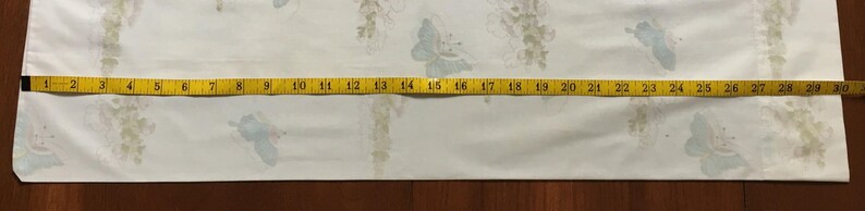 May include: A white fabric with a floral pattern and blue butterfly accents. A yellow measuring tape shows a length of approximately 30 inches. The fabric is likely used for sewing or crafting projects.
