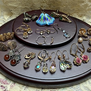 May include: A collection of assorted vintage earrings displayed on a dark wood stand. The set includes gold-tone metal pieces, colorful enamel floral designs, clear crystal drops, small hoop earrings, and floral-shaped studs with faux pearls.