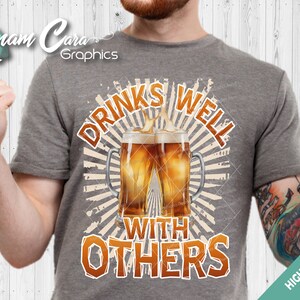 Op de afbeelding: Een grijs T-shirt met een vintage-stijl grafische afbeelding van twee bierglazen met de tekst "Drinks Well With Others".