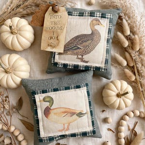 May include: Two square sage green fabric pillows featuring centered patches with illustrations of ducks. The top pillow has a tan tag with the text "INTO THE WOODS I GO!" and a small metal bell. The bottom pillow displays a mallard duck.