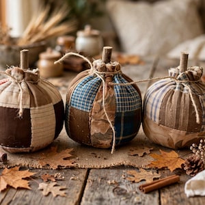 May include: Three fabric pumpkins crafted from various patches of plaid, checkered, and solid-colored textiles in shades of brown, blue, and cream. Each pumpkin features a short wooden stem and is tied with a thin piece of natural twine.