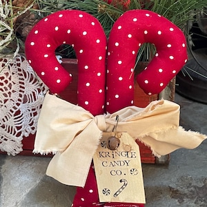 May include: Two red fabric candy canes with white polka dots, tied together with a cream-colored bow. A tag reads "Kringle Candy Co." with a candy cane illustration. Festive holiday decor.