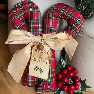 May include: Two fabric candy canes featuring a red, green, and yellow plaid pattern. A beige frayed ribbon is tied around the center, holding a rectangular paper tag with the text MERRY & BRIGHT and two holly leaf icons, plus a small metal bell.