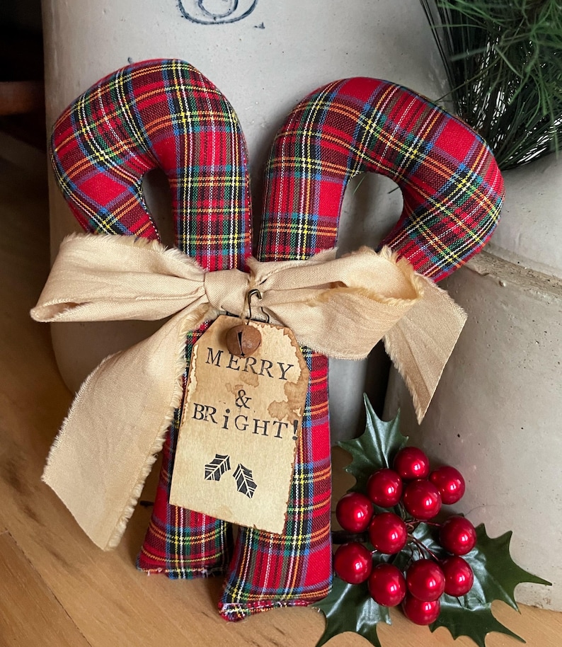 May include: Two fabric candy canes featuring a red, green, and yellow plaid pattern. A beige frayed ribbon is tied around the center, holding a rectangular paper tag with the text MERRY & BRIGHT and two holly leaf icons, plus a small metal bell.