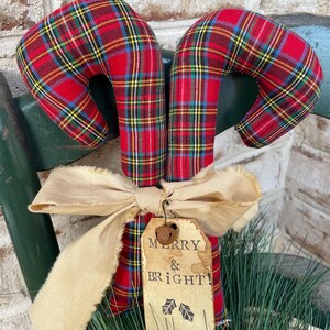 May include: Two fabric candy canes featuring a red, yellow, blue, and green plaid pattern. A beige frayed ribbon is tied around the center, holding a rectangular paper tag with the text MERRY & BRIGHT and two small leaf icons.