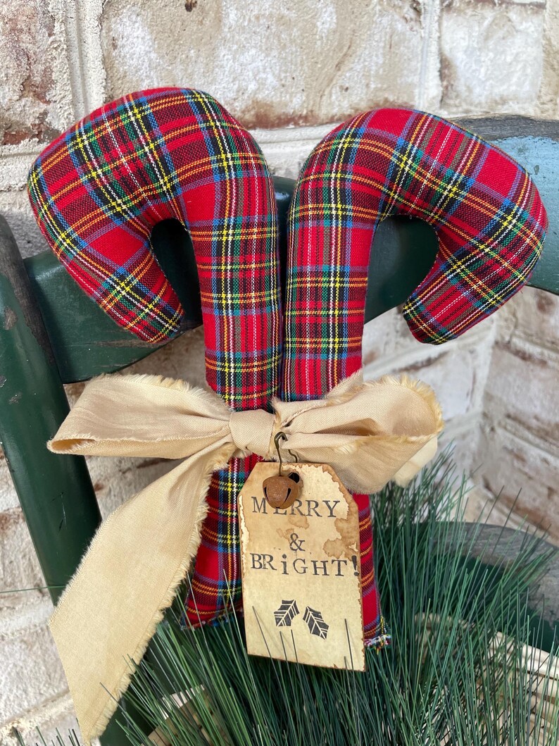 May include: Two fabric candy canes featuring a red, yellow, blue, and green plaid pattern. A beige frayed ribbon is tied around the center, holding a rectangular paper tag with the text MERRY & BRIGHT and two small leaf icons.