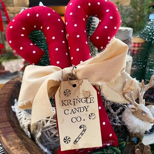 May include: Two red fabric candy canes with white polka dots, tied with a cream-colored bow. A tag reads "Kringle Candy Co." with a candy cane illustration. Festive holiday decor.