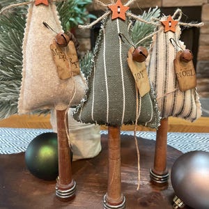 May include: Three miniature Christmas trees made of fabric with burlap ribbon and wooden bases. The trees are decorated with small bells, stars, and tags that say "Merry", "Joy", and "Noel".