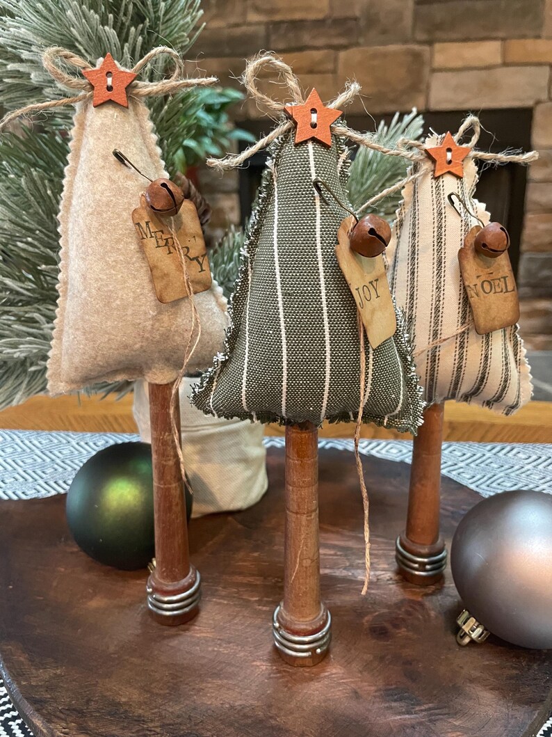 May include: Three miniature Christmas trees made of fabric with burlap ribbon and wooden bases. The trees are decorated with small bells, stars, and tags that say "Merry", "Joy", and "Noel".