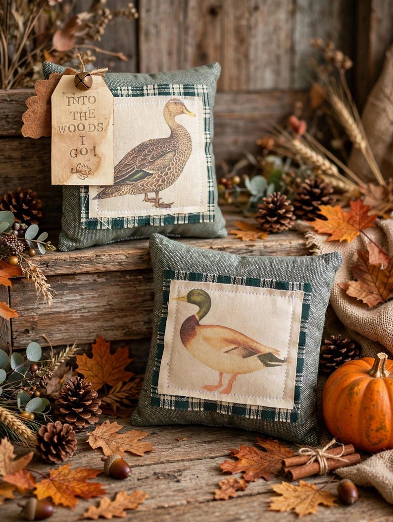 May include: Two square decorative pillows in sage green fabric with a dark green plaid border. Each features a central cream-colored patch with a duck illustration. One pillow has a tan paper tag attached with the text INTO THE WOODS I GO.