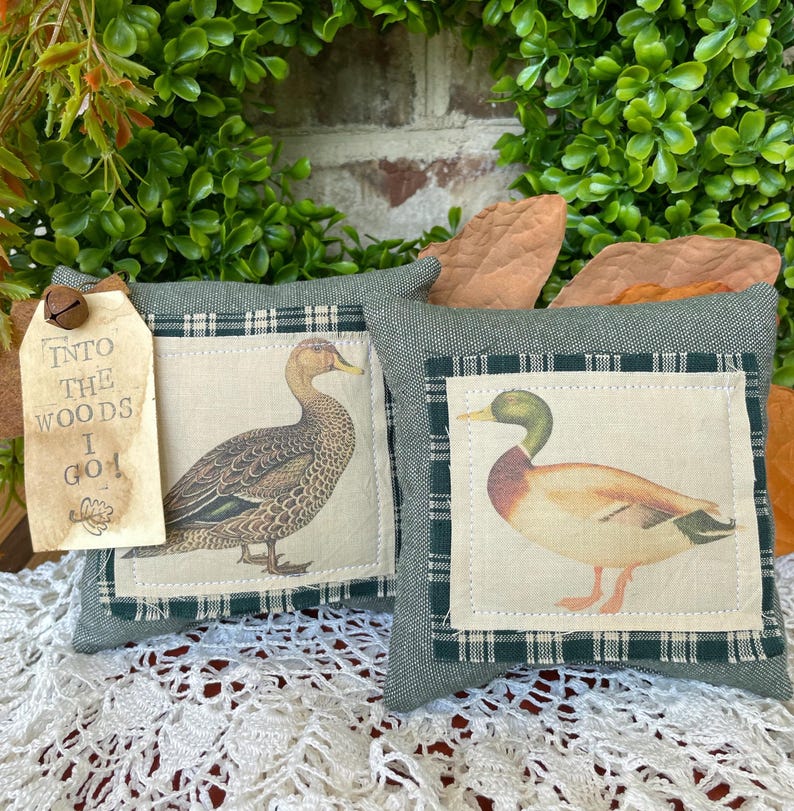May include: Two square fabric pillows in sage green with a dark green and white checkered border. Each pillow features a central panel with a duck illustration. A beige paper tag attached to the left pillow reads, INTO THE WOODS I GO!