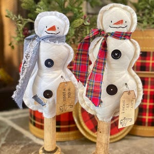 May include: Two handmade snowman ornaments. Each snowman has a white body, a carrot nose, and a black button. One wears a gray scarf, the other a red plaid scarf. Tags read "Warm Wishes" and "Let it Snow!"