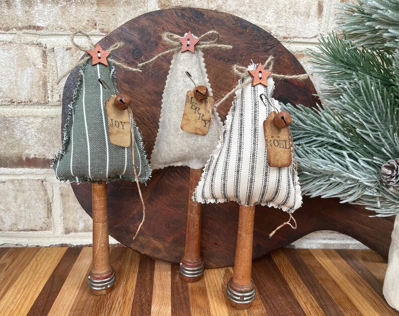 May include: Three handmade fabric Christmas trees with striped fabric and wooden spools. The trees are decorated with small bells and tags that say "Joy", "Merry", and "Noel".