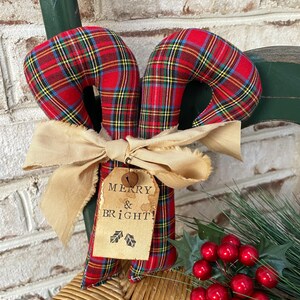 May include: Two fabric candy canes featuring a red, yellow, and blue plaid pattern. A beige frayed ribbon is tied around the center, holding a tan paper tag with the printed text MERRY & BRIGHT! and two small holly leaf icons, plus a small bell.