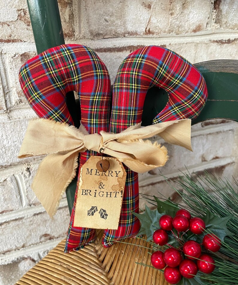 May include: Two fabric candy canes featuring a red, yellow, and blue plaid pattern. A beige frayed ribbon is tied around the center, holding a tan paper tag with the printed text MERRY & BRIGHT! and two small holly leaf icons, plus a small bell.