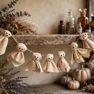 May include: A garland featuring seven small ghost figures made from light beige fabric with frayed edges. Each ghost has a rounded head with three small black dots representing eyes and a mouth, all strung together on a thin piece of natural twine.