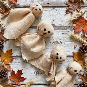 May include: A garland of four ghost-shaped ornaments made from light beige fabric with frayed edges. Each ghost features a rounded head with three small black dots forming eyes and a mouth, all strung together on a thin piece of natural twine.