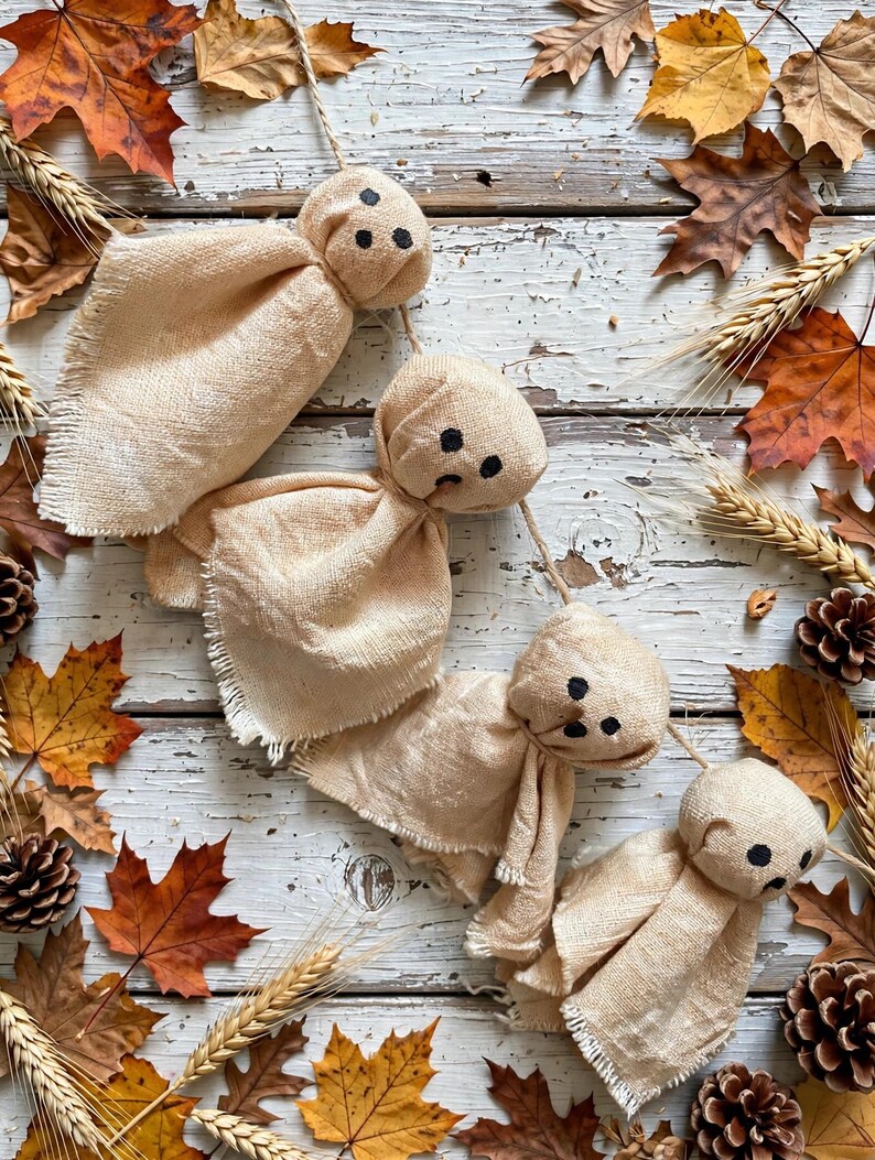 May include: A garland of four ghost-shaped ornaments made from light beige fabric with frayed edges. Each ghost features a rounded head with three small black dots forming eyes and a mouth, all strung together on a thin piece of natural twine.