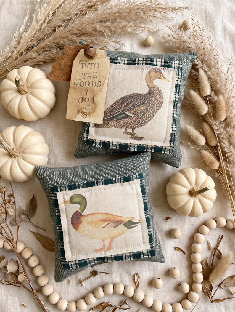 May include: Two square sage green fabric pillows featuring centered patches with illustrations of ducks. The top pillow has a tan tag with the text "INTO THE WOODS I GO!" and a small metal bell. The bottom pillow displays a mallard duck.