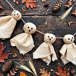 May include: A garland featuring four ghost-shaped figures made from light beige fabric with frayed edges. Each ghost has two black circular eyes and a small black circular mouth, all suspended from a single piece of natural-colored twine.